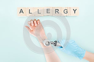 Cropped view of doctor examining hand of woman with dermatoscope near wooden blocks with word allergy isolated on blue.