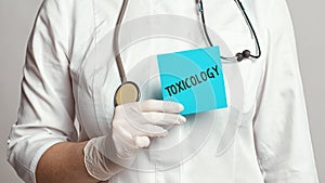 Cropped view of doctor in a white coat and sterile gloves holding a note with text - Toxicology