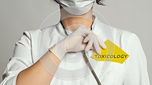 Cropped view of doctor in a white coat and sterile gloves holding a note with text - Toxicology