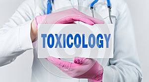 Cropped view of female doctor in a white coat and pink sterile gloves holding a card with word Toxicology