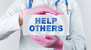 Cropped view of female doctor in a white coat and pink sterile gloves holding a card with words - Help Others. Medical concept