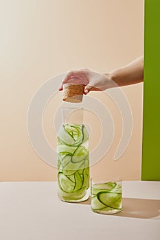 Cropped view of female hand holding cork under bottle filled with water and sliced cucumbers on table