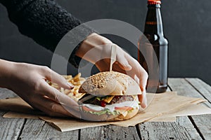 cropped view of female hands with hamburger, french fries and bottle of beer on baking paper