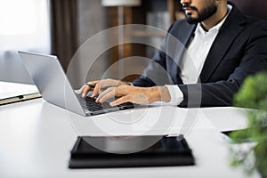 Cropped view of hands of young bearded businessman typing on laptop during work in office.