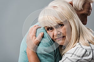 cropped view of husband hugging sad retired wife with grey hair isolated on grey.