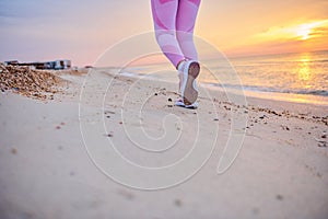 Cropped view of legs in sports shoes of sporty woman running along sea coast against background of beautiful sunrise