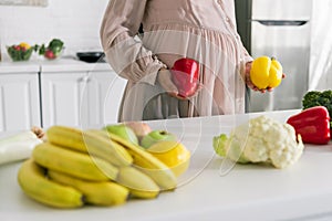 View of pregnant woman holding paprika standing near fruits on table