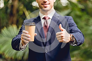 Cropped view of smiling businessman in suit and tie holding coffee to go and showing thumb up in orangery.