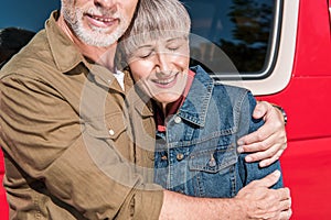 cropped view of smiling senior couple of tourists standing near red car and embracing in sunny day.