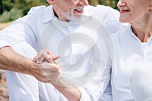 cropped view of smiling senior couple in white shirts holding hands.