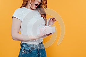 Cropped view of surprised redhead girl holding gift box isolated on yellow with copy space.