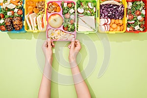 Cropped view of woman choosing lunch boxes with food on green background.
