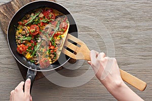 Cropped view of woman cooking omelet with mushrooms, tomatoes and greens on frying pan with wooden shovel.