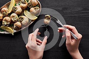 Cropped view of woman eating delicious escargots with lemon on black wooden table.