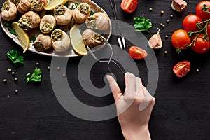 Cropped view of woman eating delicious escargots with lemon on black wooden table.