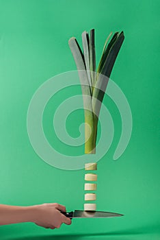 Cropped view of woman holding knife with green sliced leek on green background.