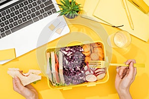Cropped view of woman holding sandwich in hand near lunch box with food, laptop, glass of water and office supplies.