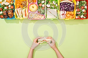 Cropped view of woman holding sandwich in hands near lunch boxes with food.