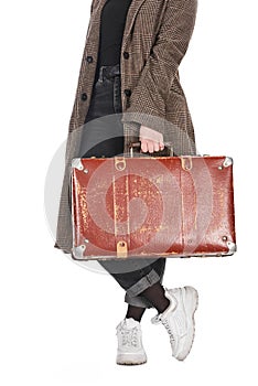 Cropped view of woman in plaid coat with crossed legs holding vintage weathered suitcase isolated on white.