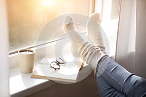 Cropped view of woman putting her feet in warm comfy socks on windowsill with book and coffee, closeup