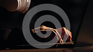 Cropped view of young computer gamer in glasses with headphones on neck typing on laptop keyboard, plays pc games