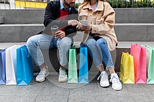 Cropped view of young diverse couple with gift bags and smartphone sitting on stairs, shopping online outdoors