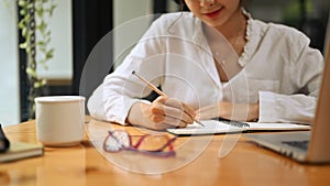 Cropped view of young woman employee making notes on notebook and working with laptop at office desk