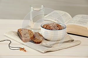 Cross, buckwheat porridge, bread, glass of water and Bible on white wooden table. Lent season