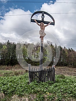 Kříž na horské louce s lesem na pozadí a modrou oblohou s mraky