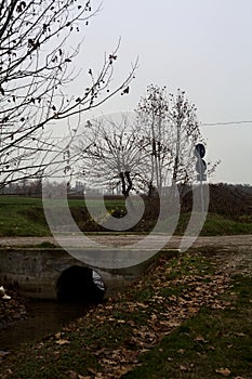 Crossing between a dirt trail and a paved one next to a stream of water on a cloudy day in the italian countryside