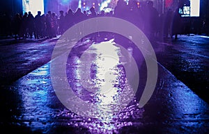 A crowd of people stands on wet pavement in front of the stage at a concert. light from event is beautifully reflected