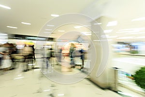 Crowd rushing inside a modern wide bright mall hall with boutiques, glass display windows, people in motion blur