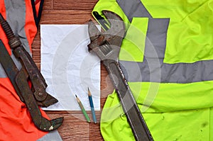 A crumpled sheet of paper with two pencils surrounded by green and orange working uniforms and adjustable wrenches. Still life as