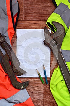 A crumpled sheet of paper with two pencils surrounded by green and orange working uniforms and adjustable wrenches. Still life as