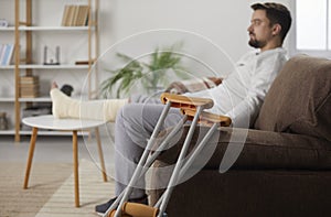 Young man with injured leg sitting at home on sofa in rehabilitation with his crutches