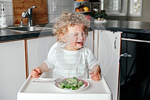 Crying Caucasian kid boy sitting in high chair with broccoli on plate. Child refusing eat healthy food. Toddler screaming in photo