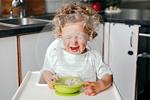 Crying Caucasian kid boy sitting in high chair with cereal puree on plate. Child refusing eat healthy food. Toddler screaming in