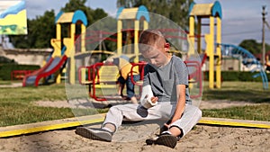 crying child with broken limb arm sits outdoors and looking how kid girl is playing on playground. concept of health