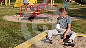 crying child with broken limb arm sits outdoors and looking how kid girl is playing on playground. concept of health