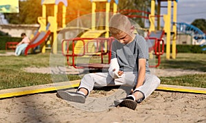 crying child with broken limb arm sits outdoors and looking how kid girl is playing on playground. concept of health