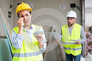 Crying construction workwoman standing at building site with banknotes