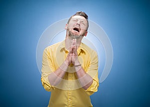 Crying disappointed young man asking for forgiveness over blue background, dresses in yellow shirt. Sad guy looking up. Guilt