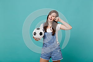 Crying disconcerted young woman football fan support favorite team with soccer ball wiping tears isolated on blue