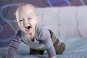 Crying happy baby boy on the bed. Laughing toddler kid looking at camera. Crawling infant in his room
