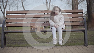 Crying lost little girl rubbing eyes as sitting on bench in spring or autumn park. Portrait of pretty lonely Caucasian