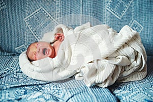 Crying newborn baby lying on a sofa, covered by a white blanket. Side view.