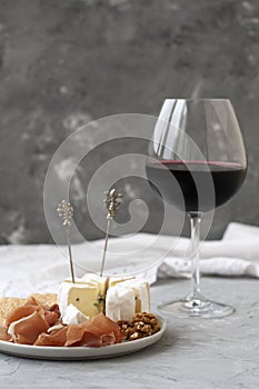 Crystal glass with red wine, in the foreground a plate with ham, nuts, crackers and Camembert cheese. Composition on a gray