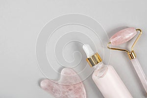 Crystal rose quartz facial roller, massage tool Gua sha and anti-aging collagen, serum in glass bottle on grey background. Facial