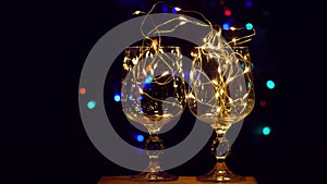 Crystal wine glasses decorated with golden glowing garland on New Year Christmas table with bokeh lights background 4K.
