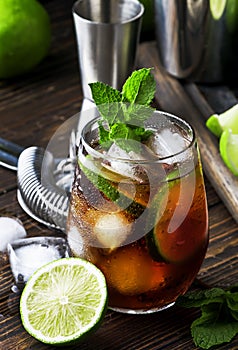 Cuba libre alcohol cocktail with golden rum, lemon juice, cola, lime and ice, dark bar counter background, bar tools, place for